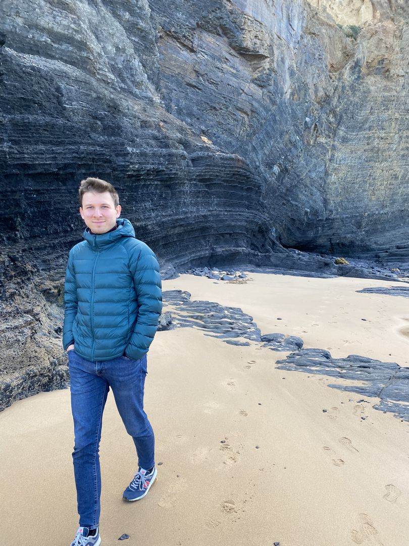 A man in a blue jacket standing on a beach near cliffs.